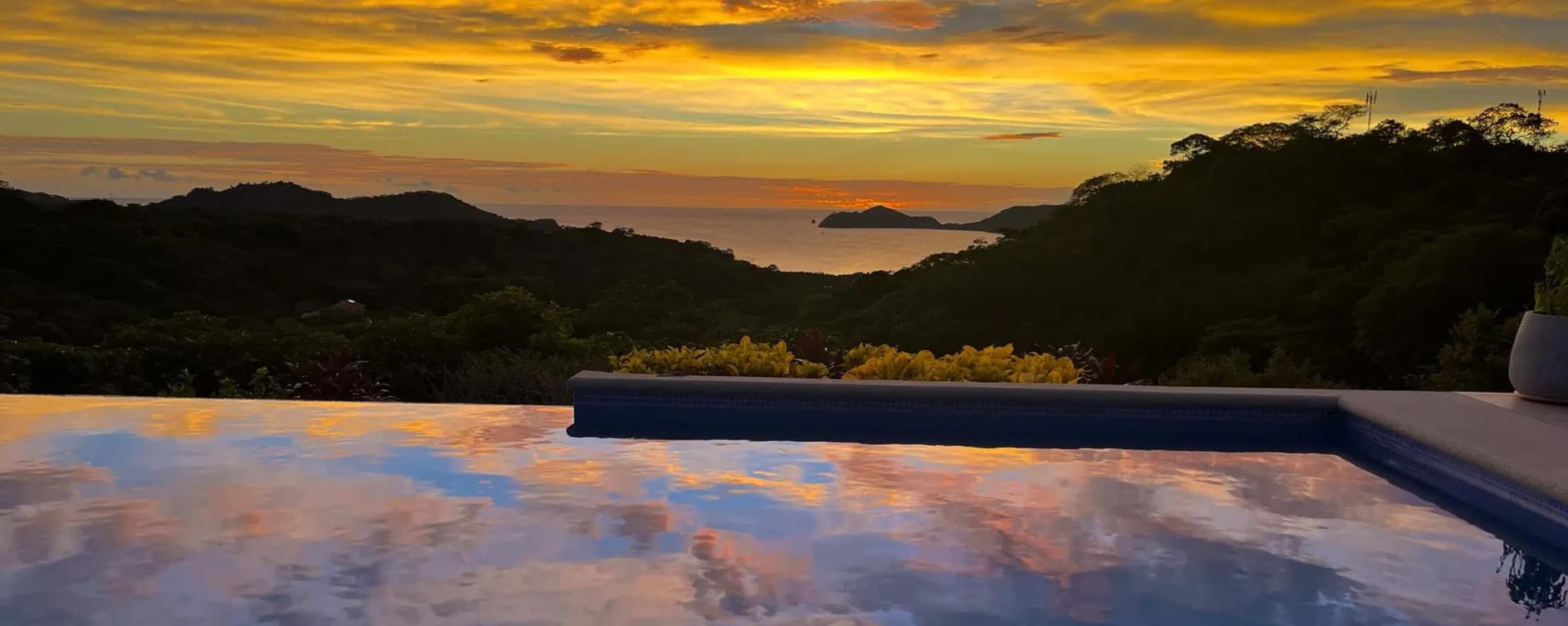 An infinity pool at sunset, reflecting an orange sky above forested hills and the sea